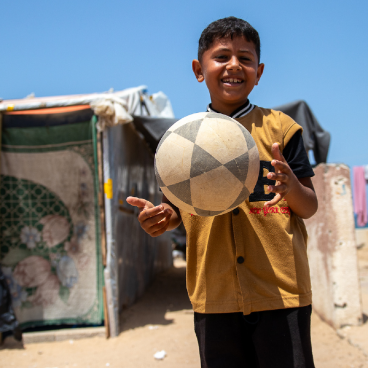Bambino sorridente in un campo rifugiati con un pallone da calcio in mano.